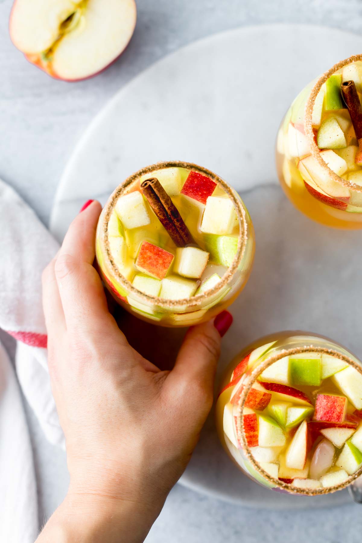 A hand holding a glass of apple cider sangria filled with diced apples and rimmed with cinnamon sugar. 