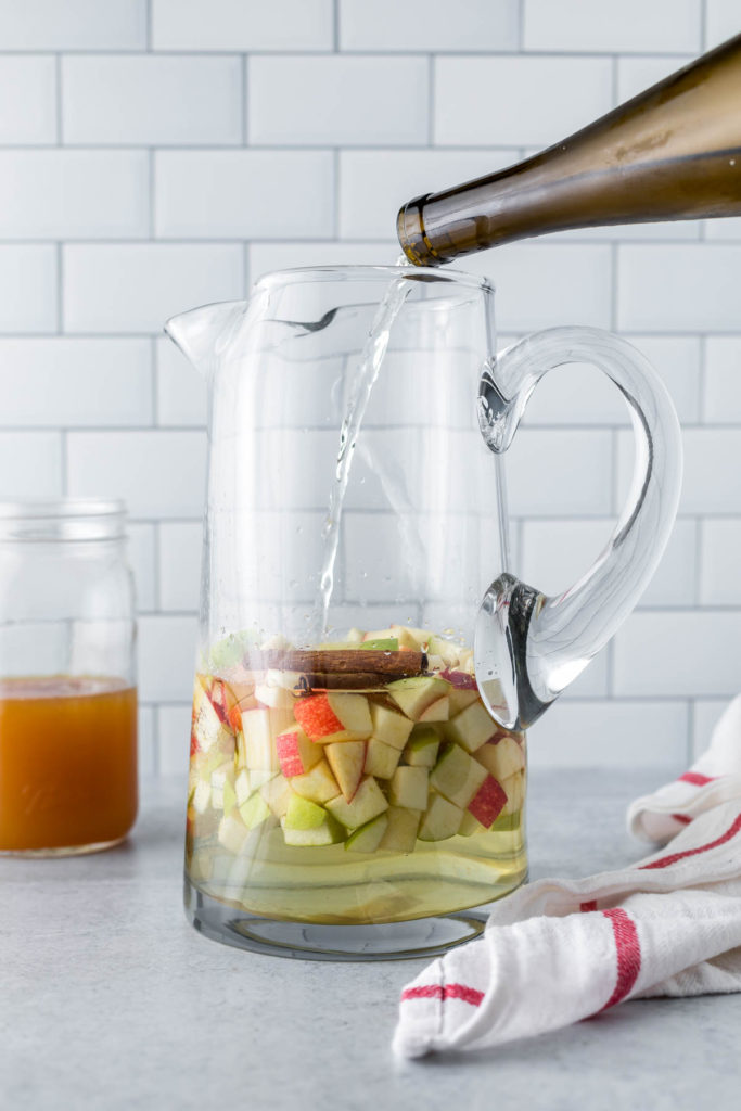 Pouring dry white wine over diced apples and cinnamon sticks in a cocktail pitcher. 
