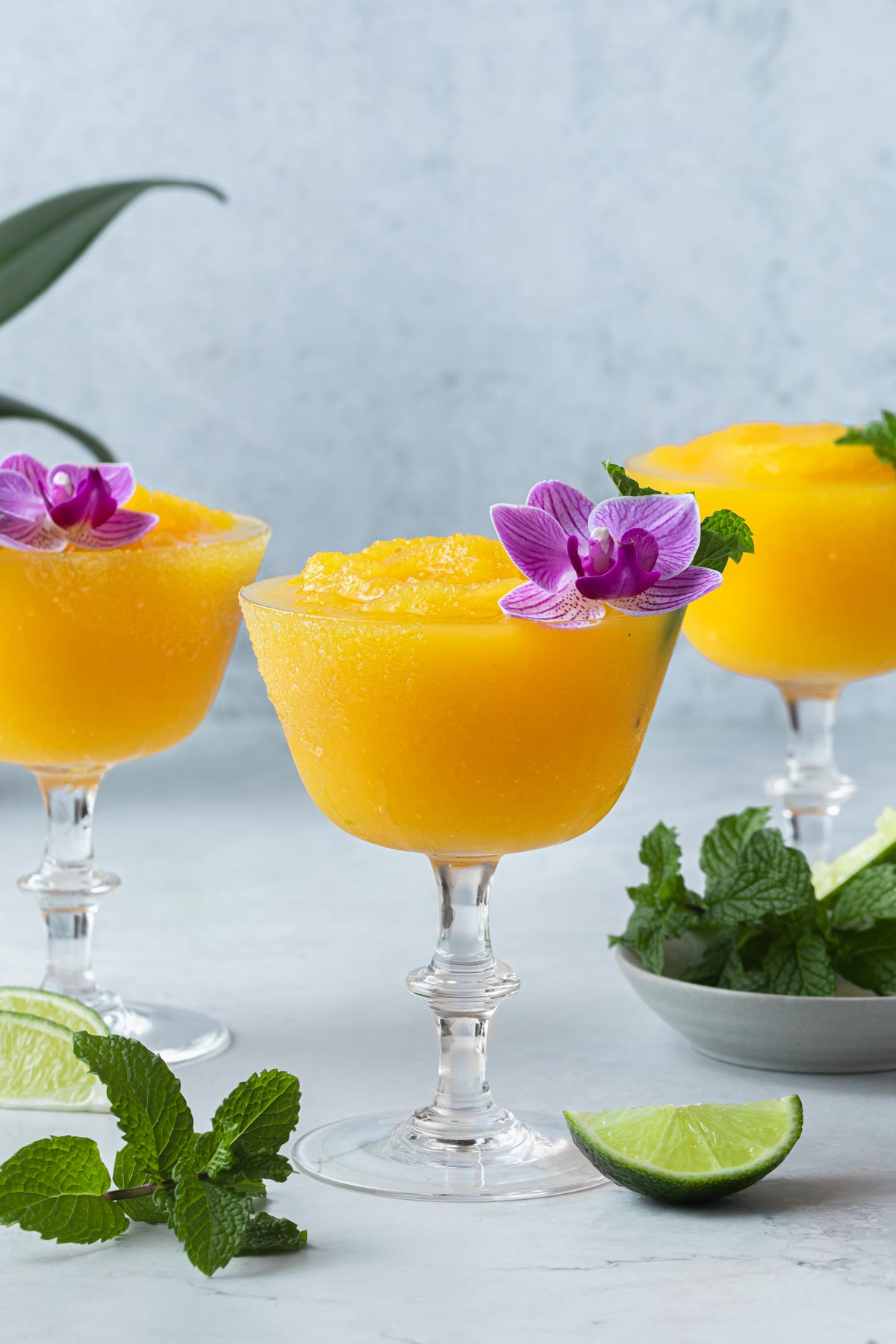  Passionfruit daiquiris in glasses garnished with an orchid and mint leaves, with a small bowl of mint leaves and wedges of lime.
