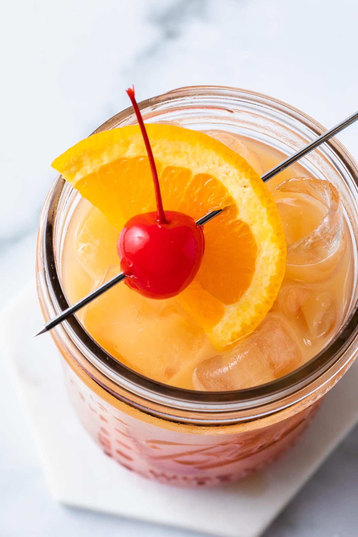 A close up of a vodka sunrise in a mason jar garnished with a maraschino cherry and an orange slice. 