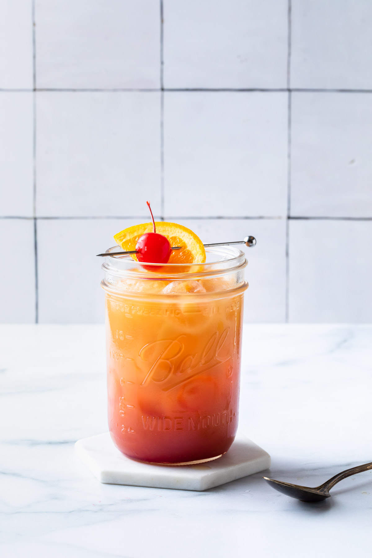 A vodka sunrise in a mason jar glass on a coaster, garnished with a maraschino cherry and an orange slice.