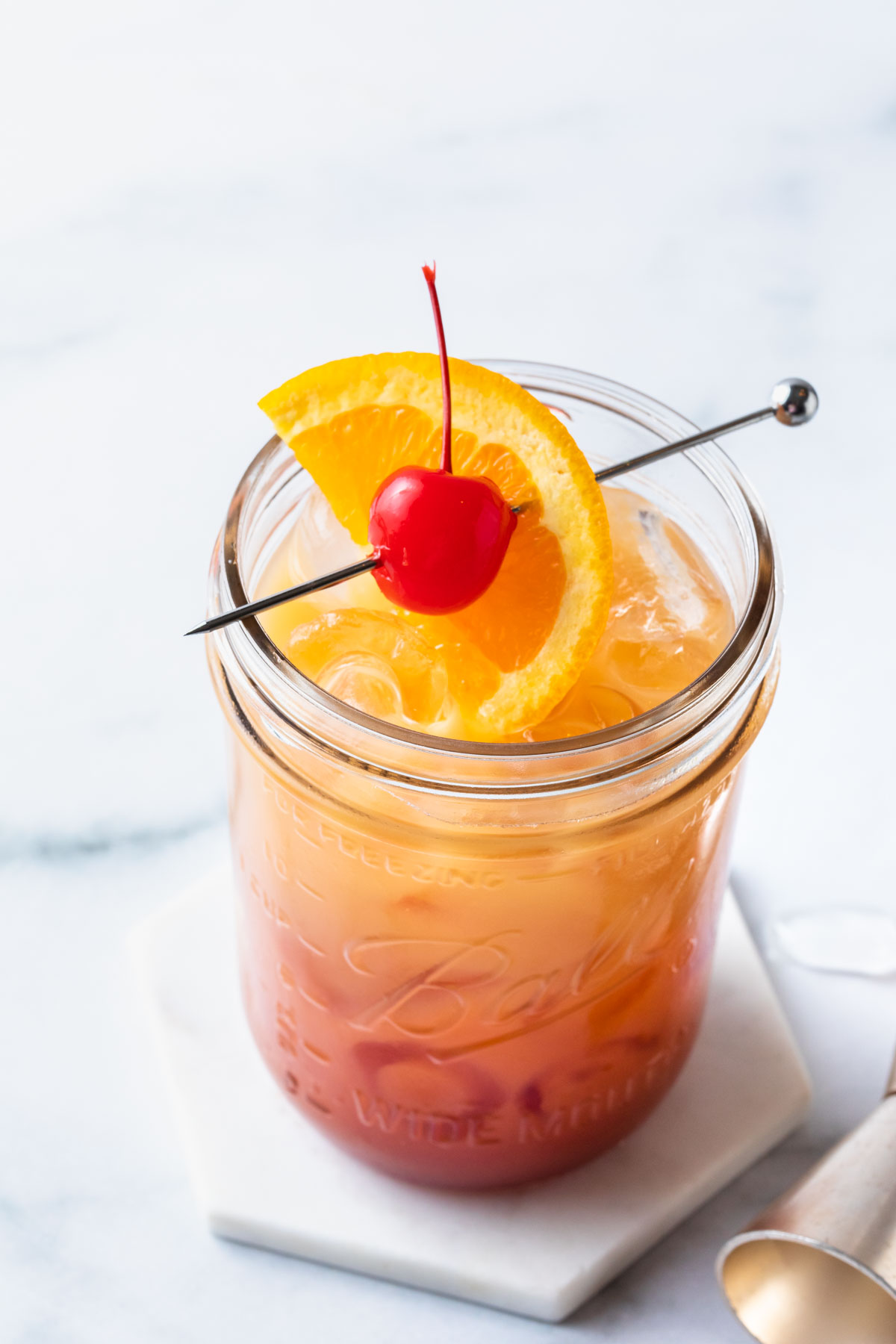 A vodka sunrise on a coaster in a mason jar glass, garnished with a maraschino cherry and an orange slice. 