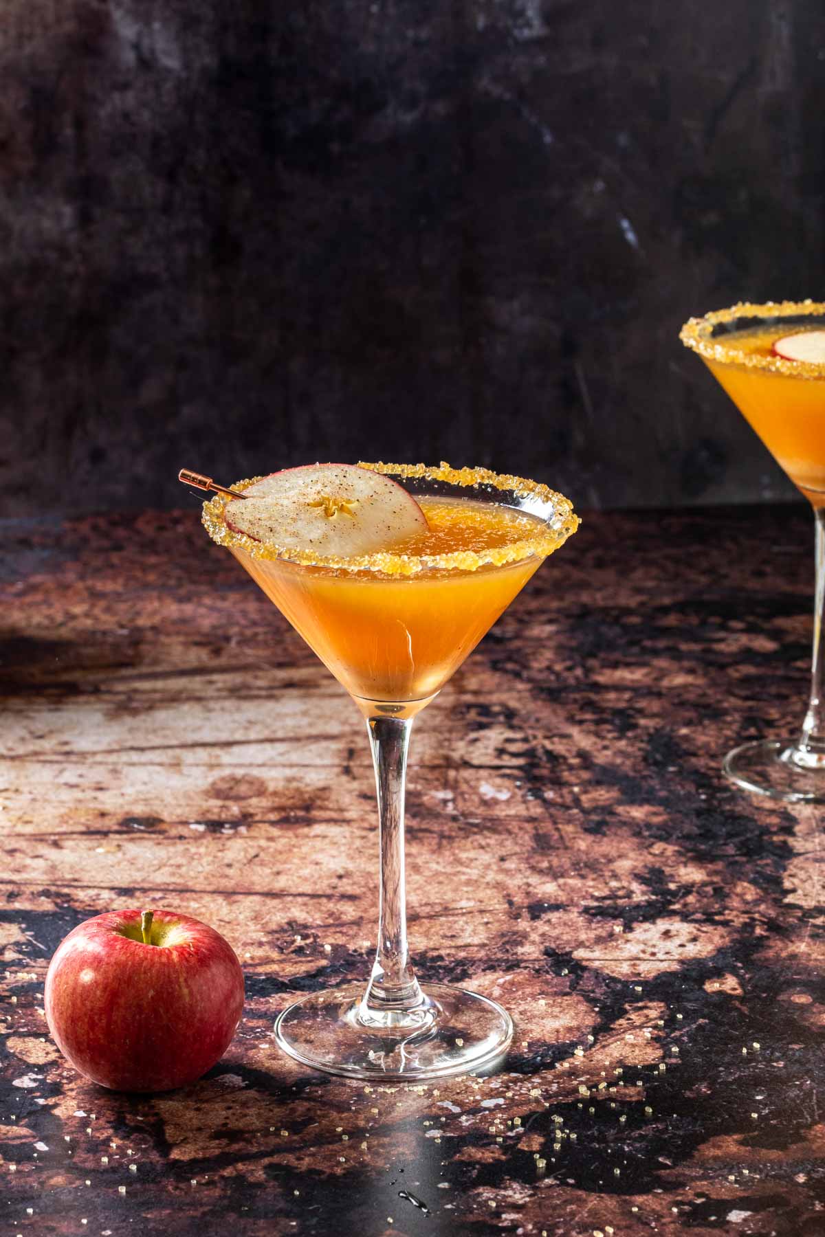 A caramel apple martini on a dark counter with an apple next to it, and another martini in the background. 