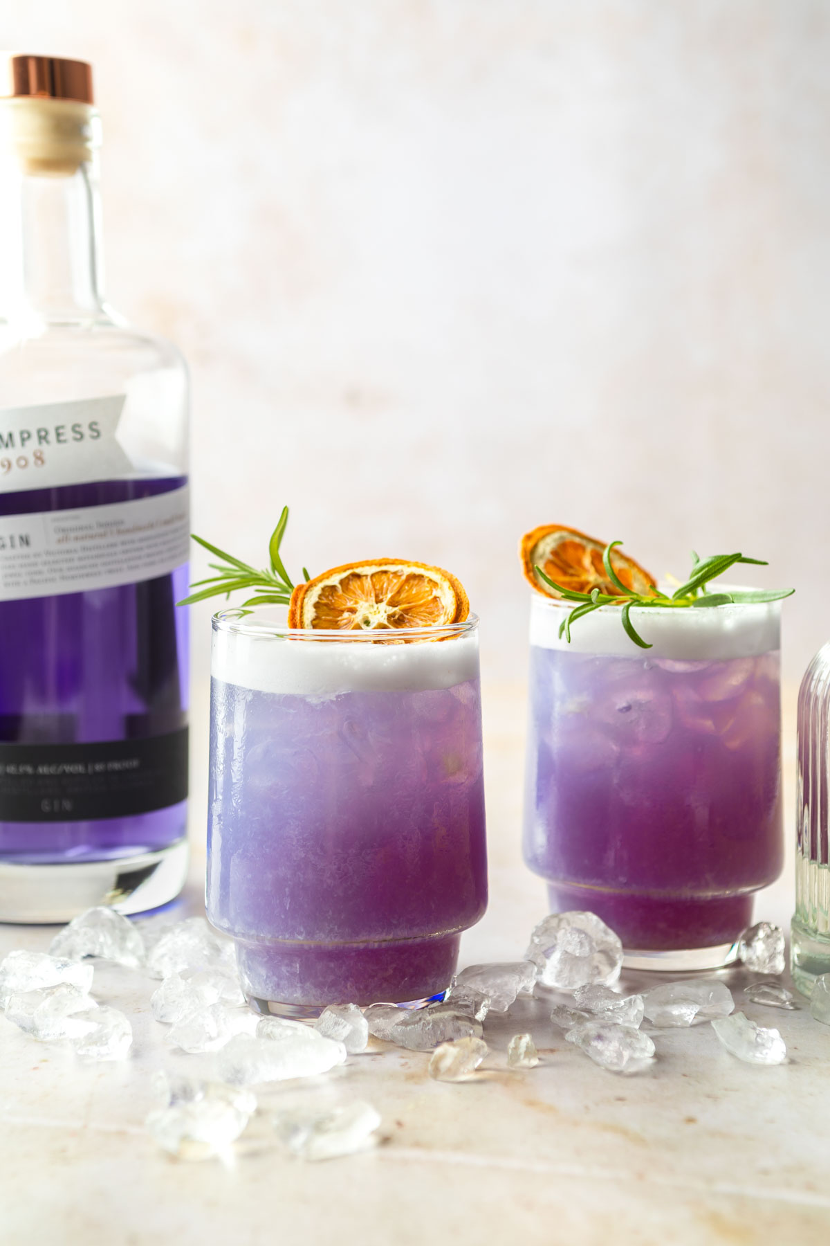 Two purple empress gin cocktails garnished with rosemary sprigs and dried orange slices, with a bottle of purple empress gin in the background.
