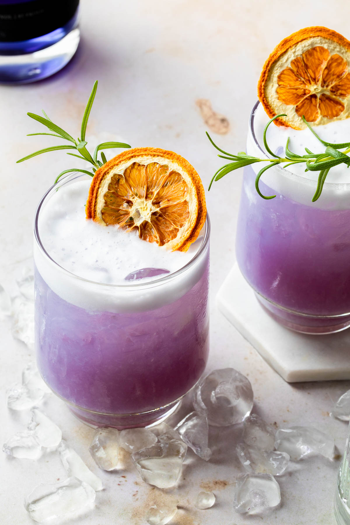 2 glasses of a purple empress gin cocktail with white foam, garnished with a dried orange and rosemary sprigs. 