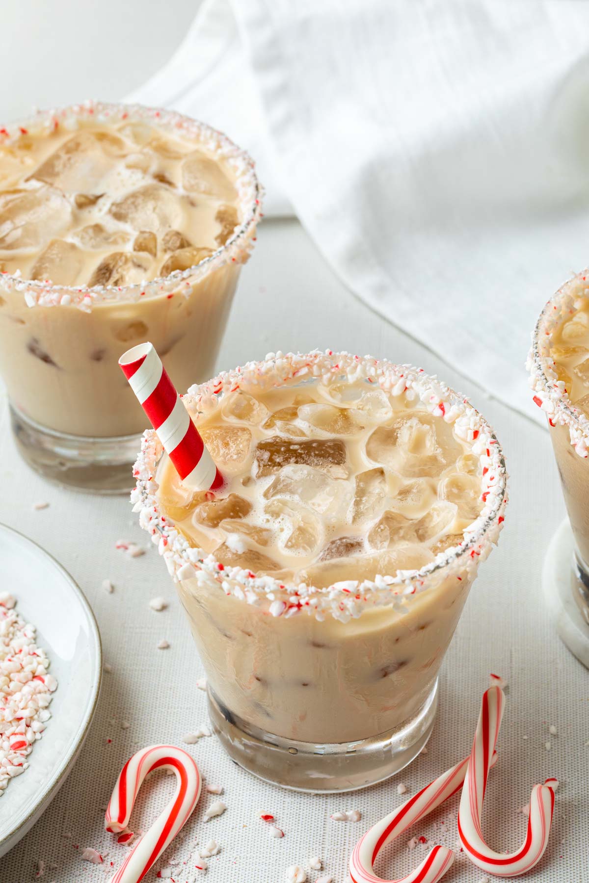 Peppermint white Russians in glasses rimmed with candy cane pieces, with red and white paper straws in the glass. 