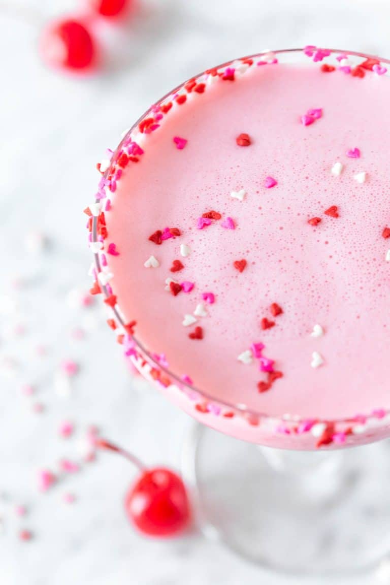 Close up overhead shot of a pink squirrel in a coupe glass garnished with pink, red and white heart sprinkles. 