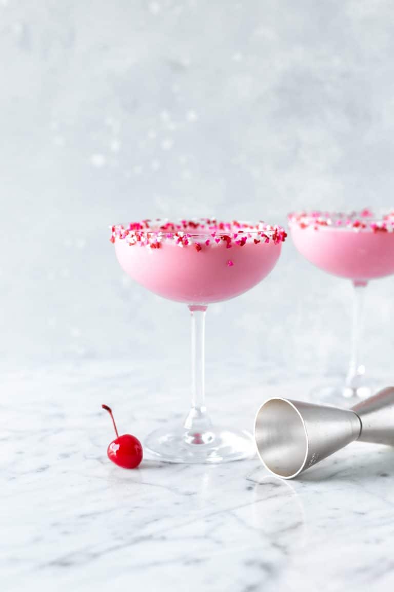 Two pink squirrel cocktails rimmed with heart sprinkles in coupe glasses on a counter with a cherry and a cocktail jigger on the counter. 