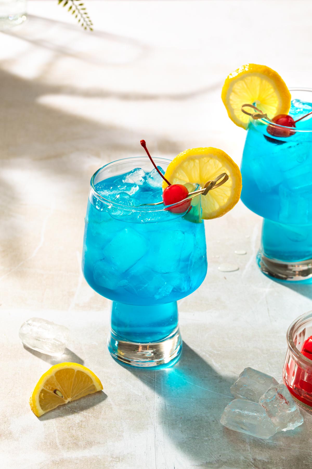 2 blue motorcycle cocktails in a glasses garnished with a lemon wheel sand a cherries. 