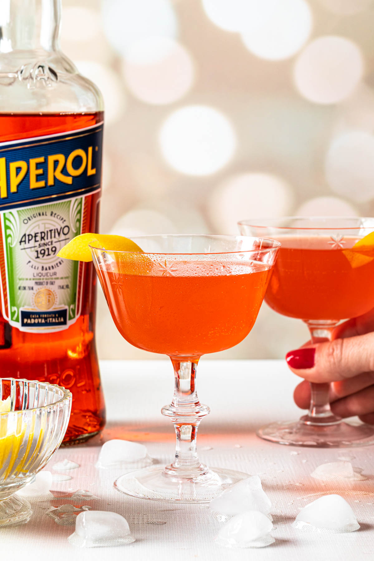 Two aperol sours with lemon twists, a bottle of aperol in the background with a hand lifting the back glass up. 