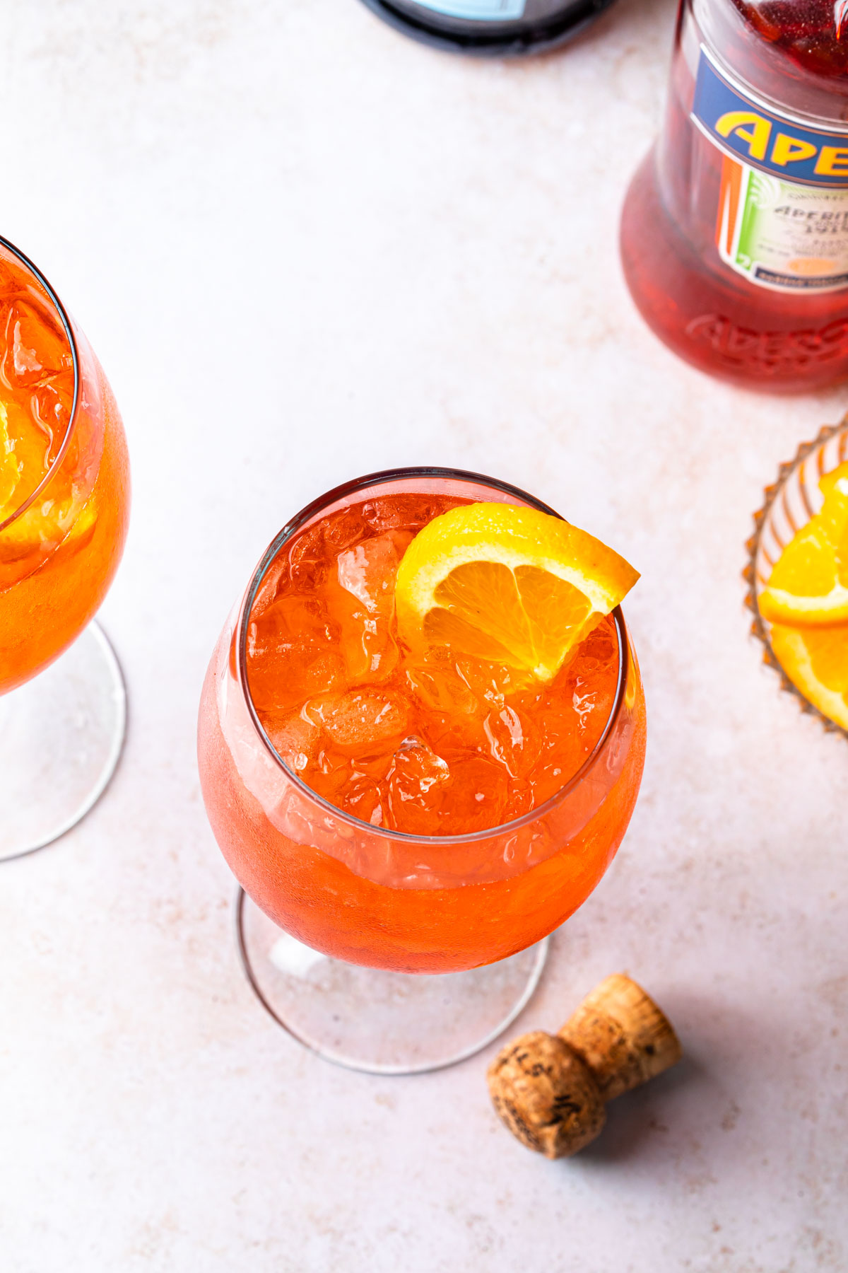 Overhead of aperol spritzer cocktails with a bowl of orange wedges and a bottle of aperol. 