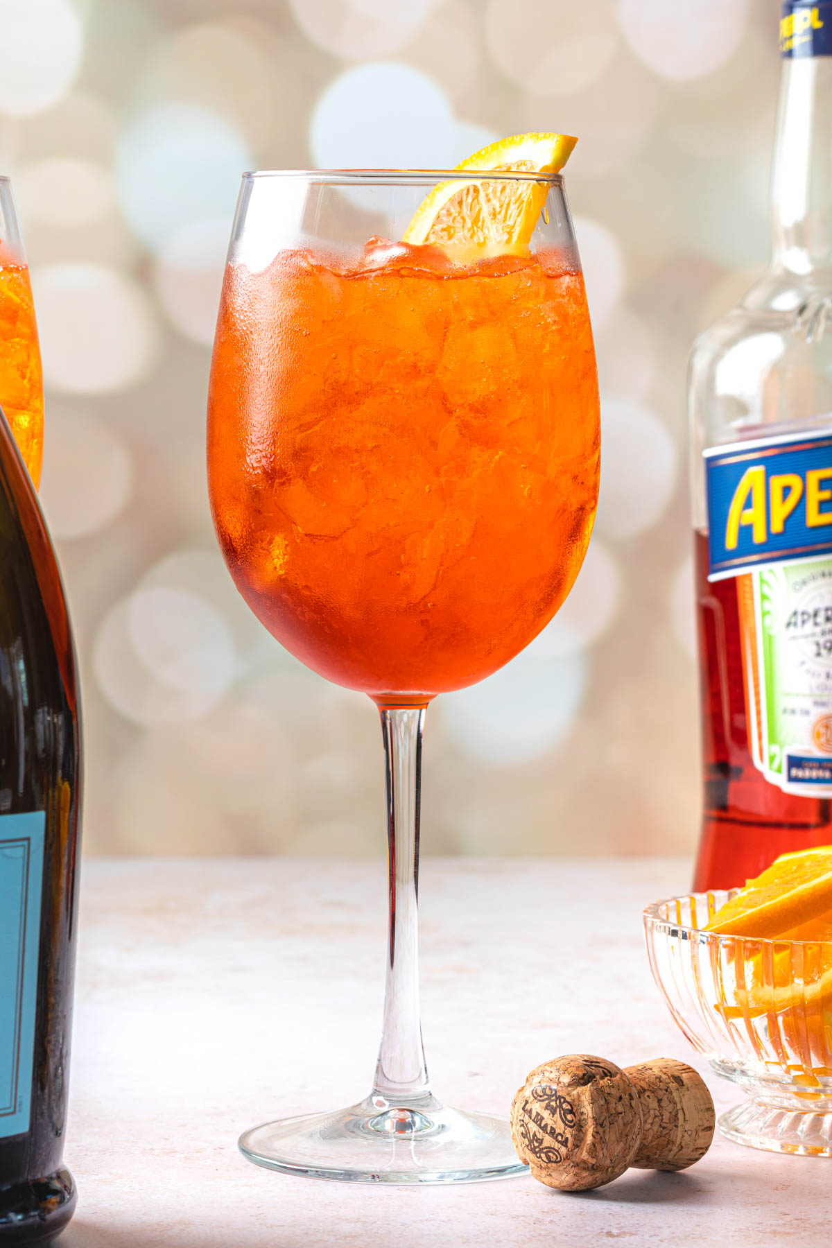 A bottle of prosecco, an aperol spritz in a wine glass with an orange wedge,  a bottle of aperol and a bowl of orange wedges. 