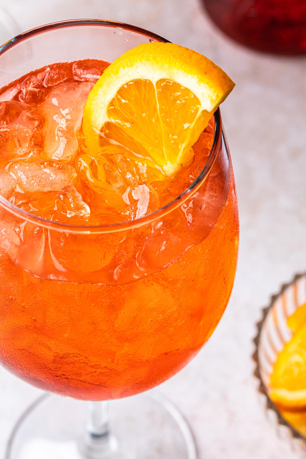A close up of an aperol spritz in a wine glass on ice with an orange wedge for garnish. 