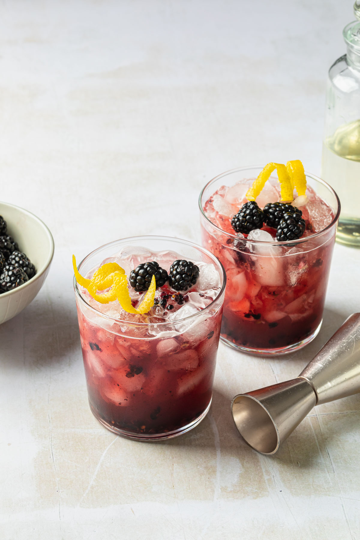 A couple of blackberry cocktails with a bowl of blackberries in the background and a cocktail jigger laying next to them. 