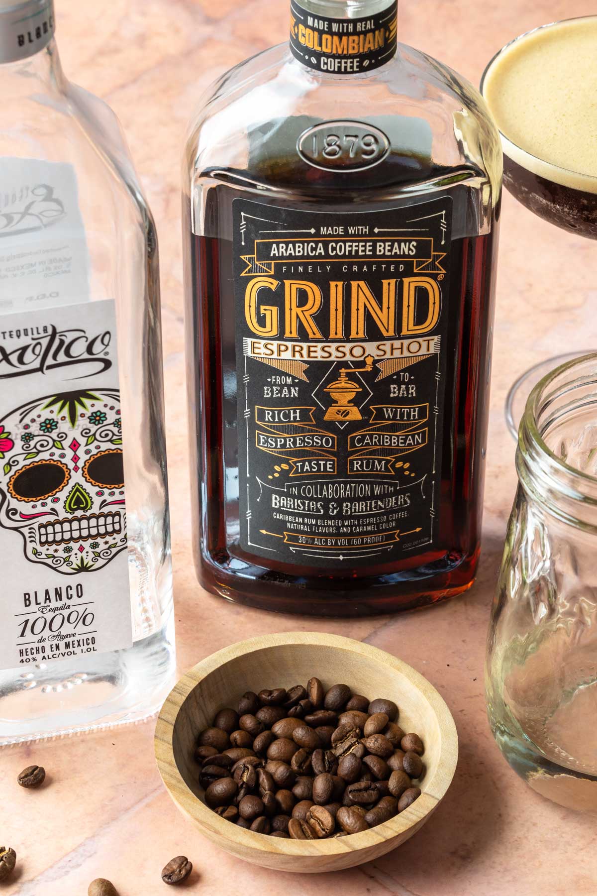 A bottle of espresso rum and tequila sitting next to a bowl of espresso beans and a bottle of simple syrup. 