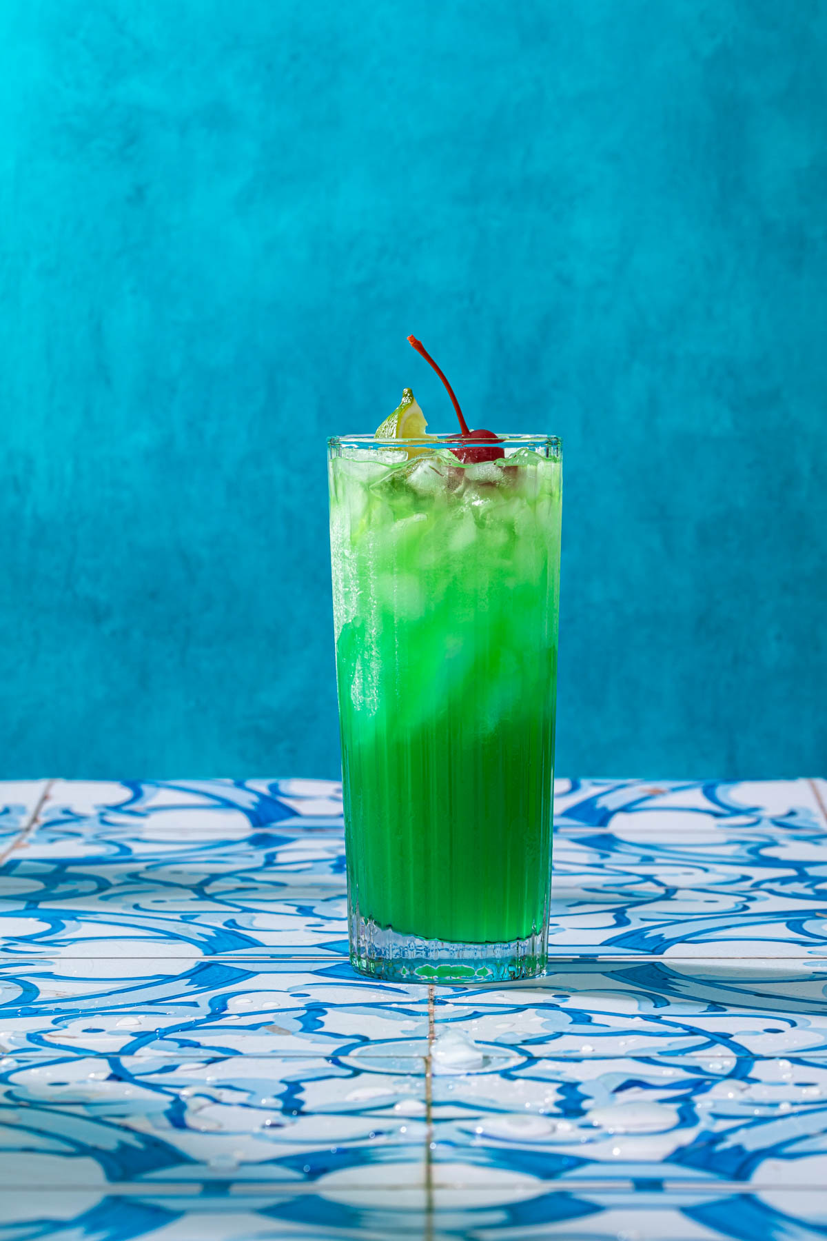 A highball glass with a bright greenish blue cocktail, garnished with a lime wedge and a maraschino cherry. 