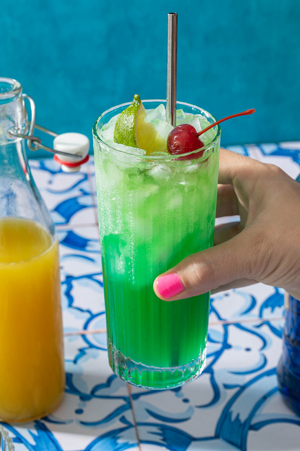 A hand holding a tropical mermaid drink, garnished with a cherry and lime wedge. 