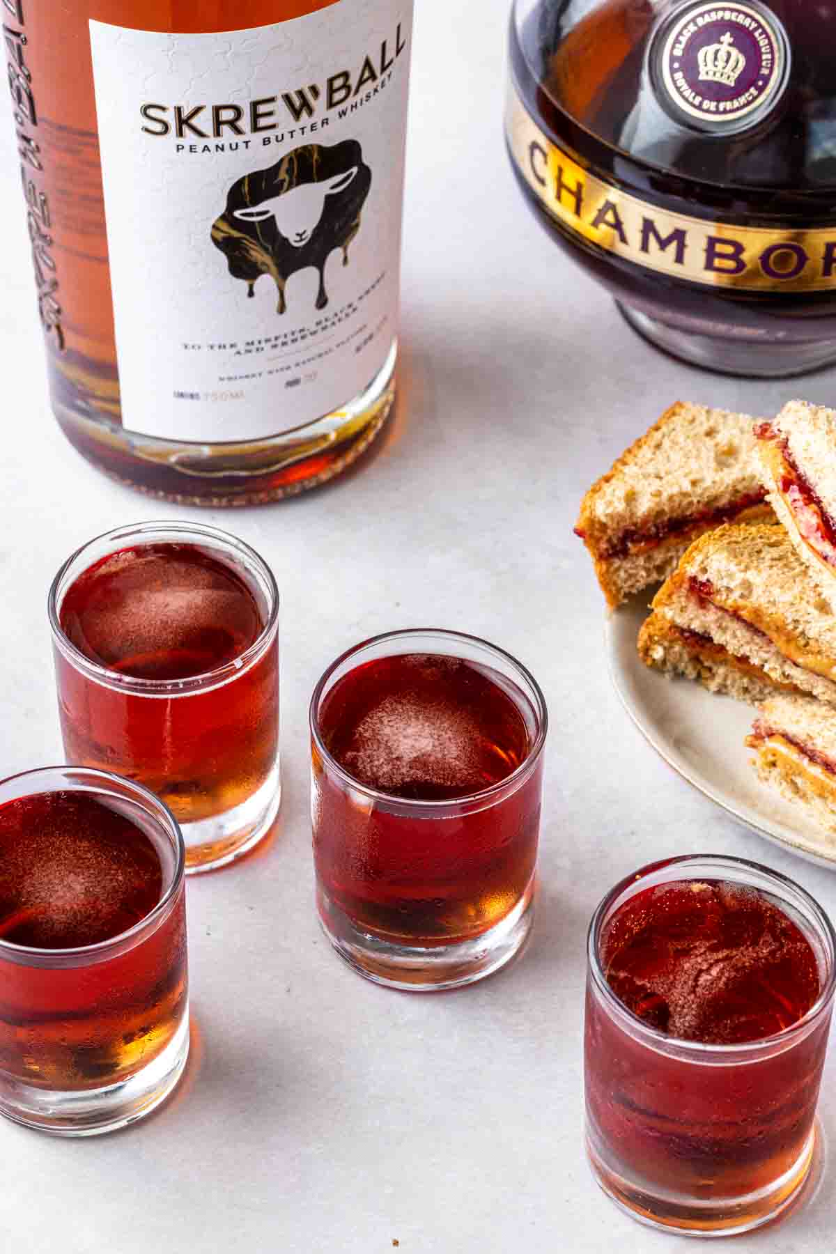 A bottle of screwball whiskey and chambord next to shots and a plate of mini pb and j sandwiches. 