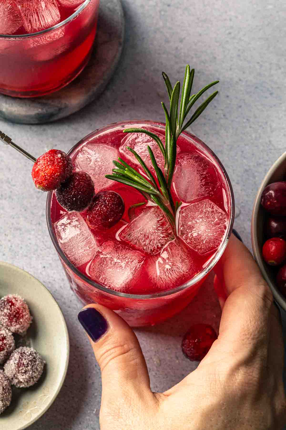 A hand holding a cranberry ginger beer holiday cocktail, garnished with rosemary and cranberries.