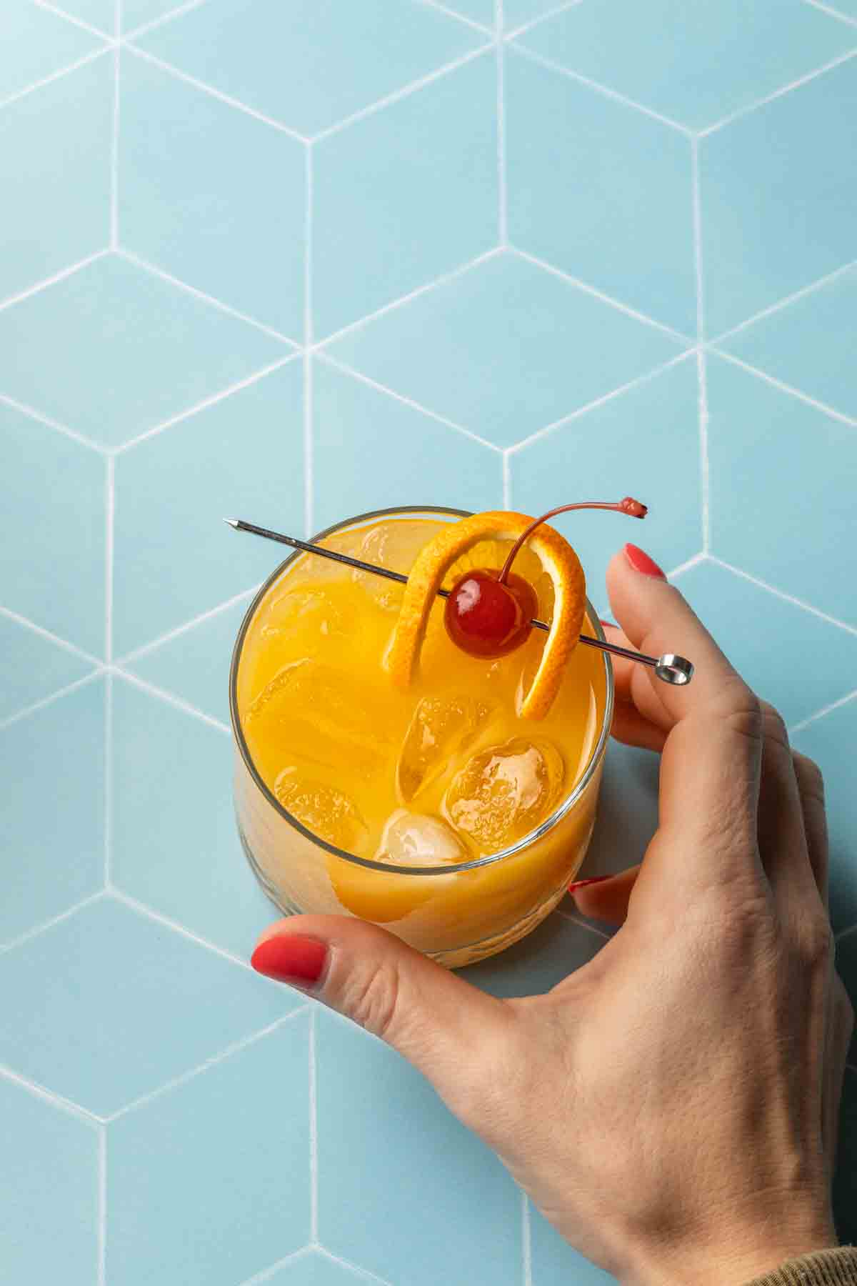 A hand holding an amaretto stone sour cocktail garnished with an orange and a cherry. 