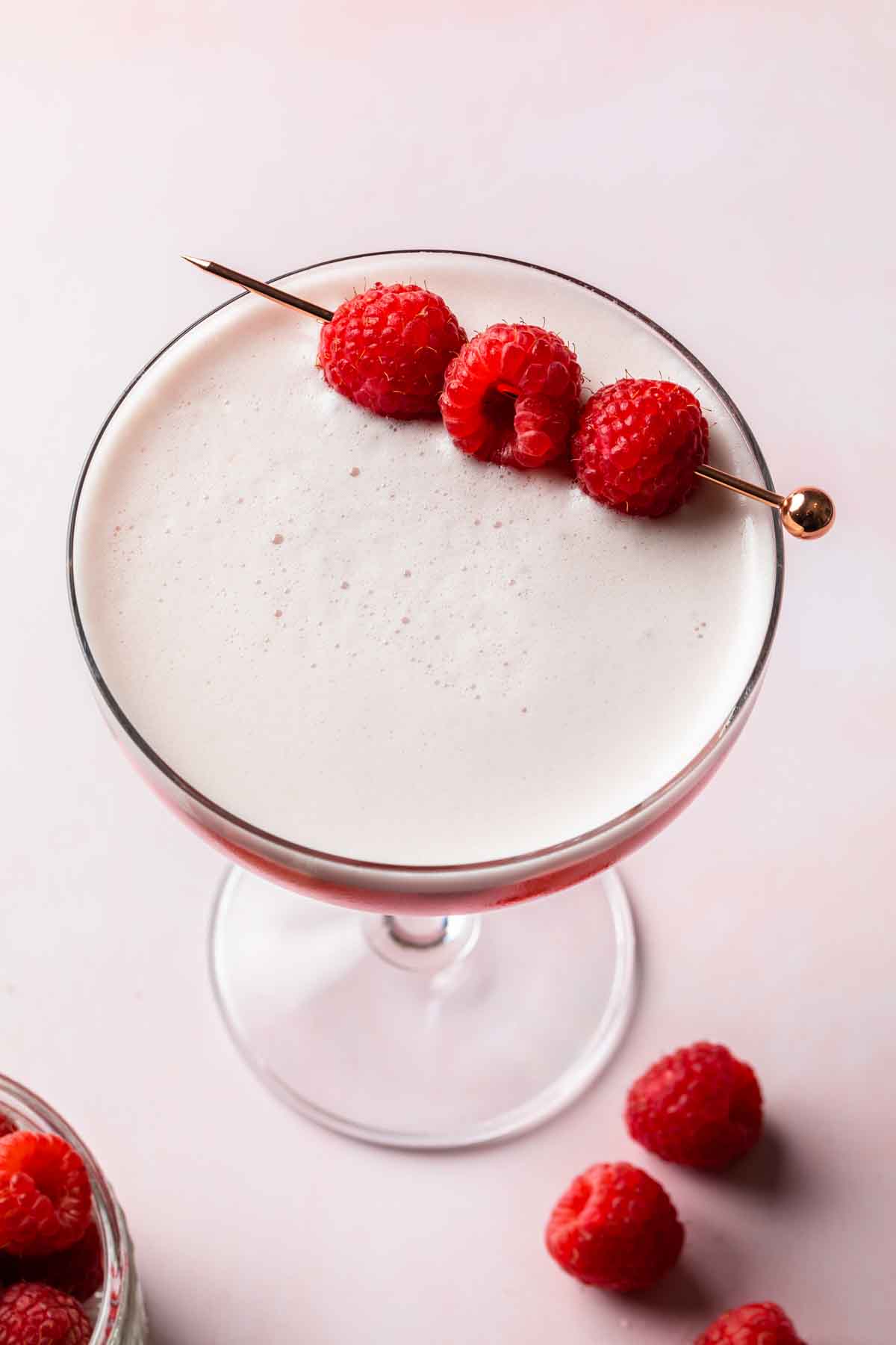 Overhead of a frothy cocktail in a coupe glass garnished with 3 raspberries. 