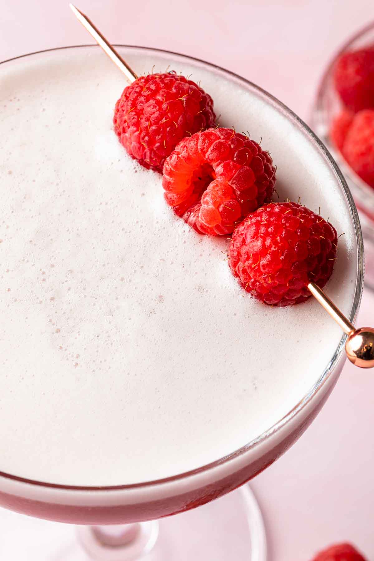 Close up of a pink cocktail with a frothy top garnished with raspberries. 