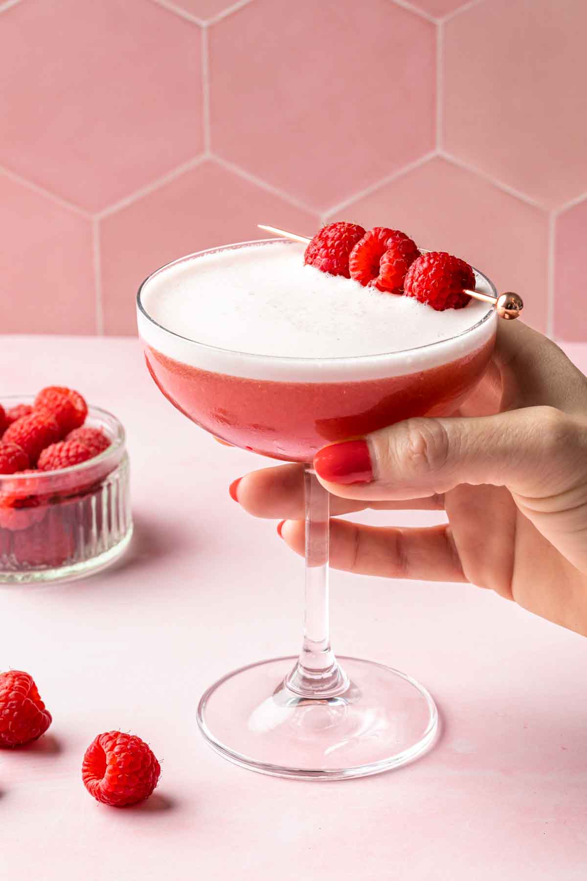 A hand holding a clover club cocktail garnished with raspberries, with a bowl of raspberries in the background. 