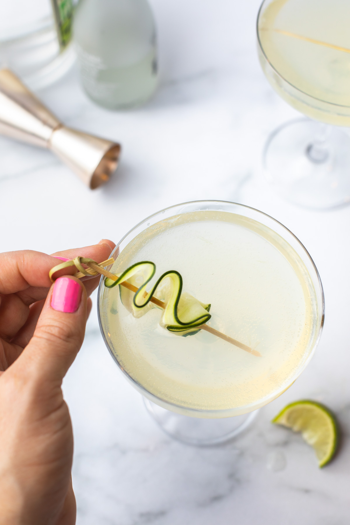 A cucumber ribbon on a toothpick being set onto a cocktail in a coupe glass. 