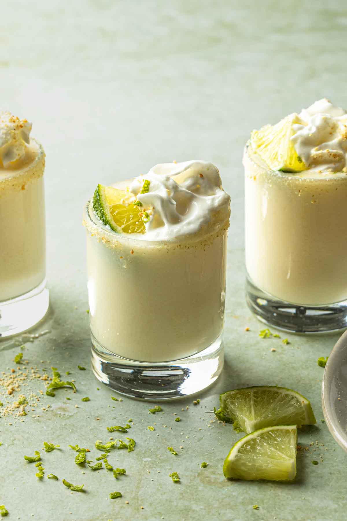 A key lime shot in a shot glass garnished with whipped cream, a lime wedge and graham cracker crumbs. 