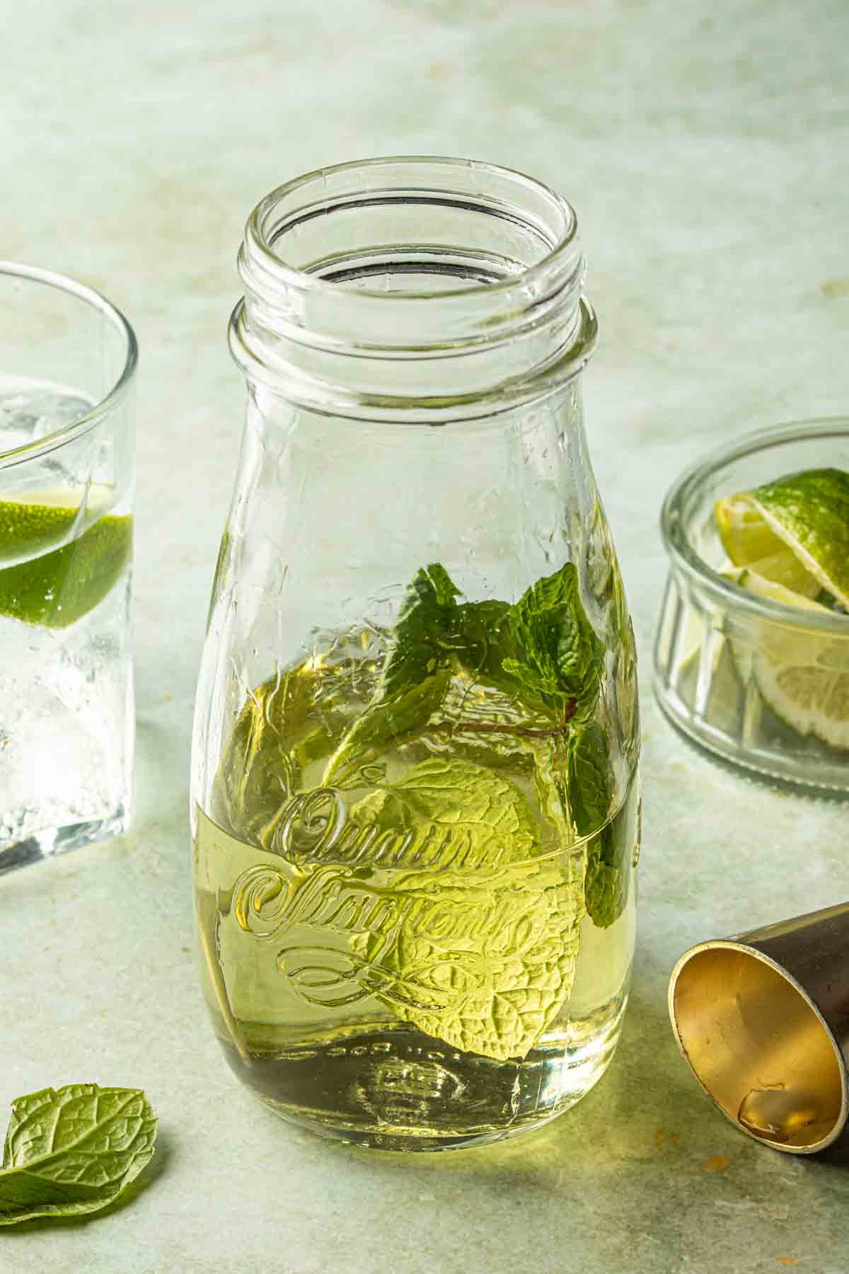 Cocktail syrup made with mint in a jar with a cocktail and a bowl of limes in the background. 