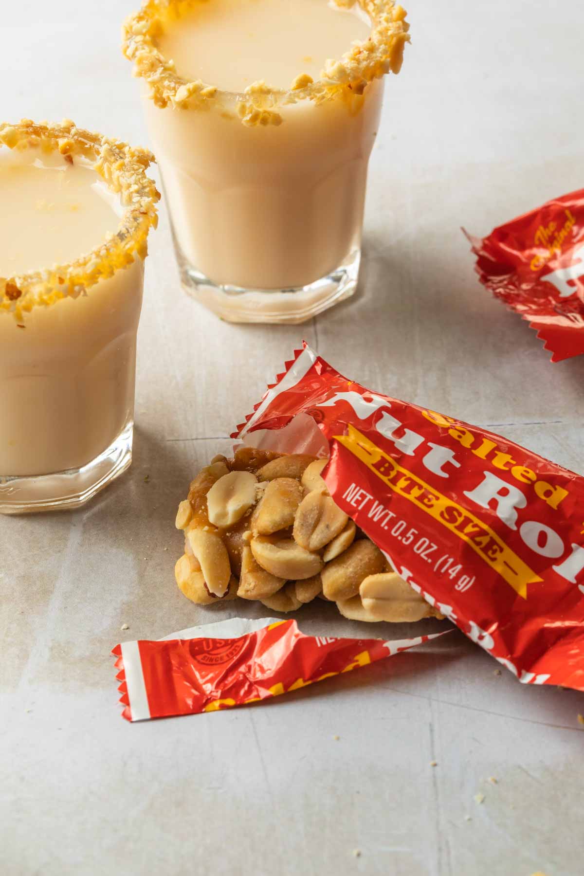 A salted nut roll candy bar open to show what is inside the package and a couple shot glasses rimmed with crushed peanuts. 