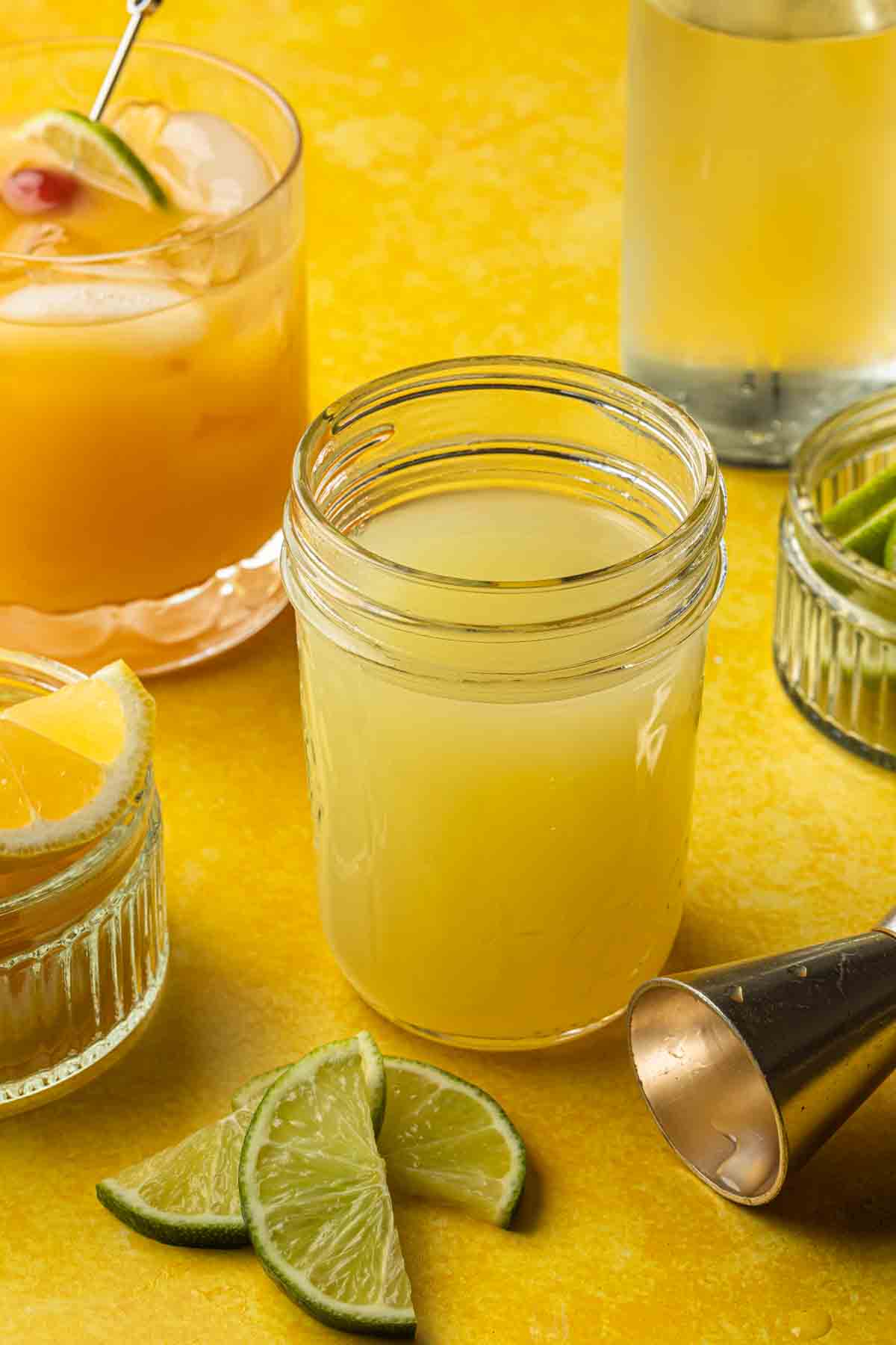 Sour cocktail mix in a jar with lemons and limes and a cocktail in the background. 