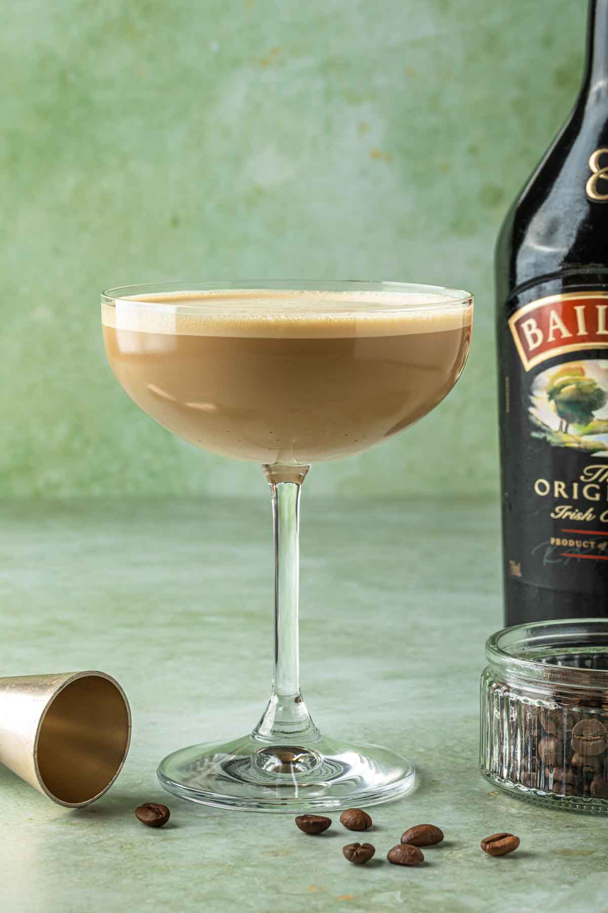 A Baileys espresso martini in a coupe glass with a bottle of baileys in the background and a bowl of espresso beans. 