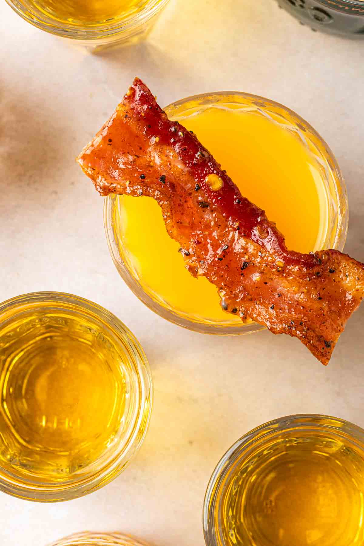An overhead shot of a glass of orange juice, topped with a piece of candied bacon next to a sweetened Irish whiskey shot. 