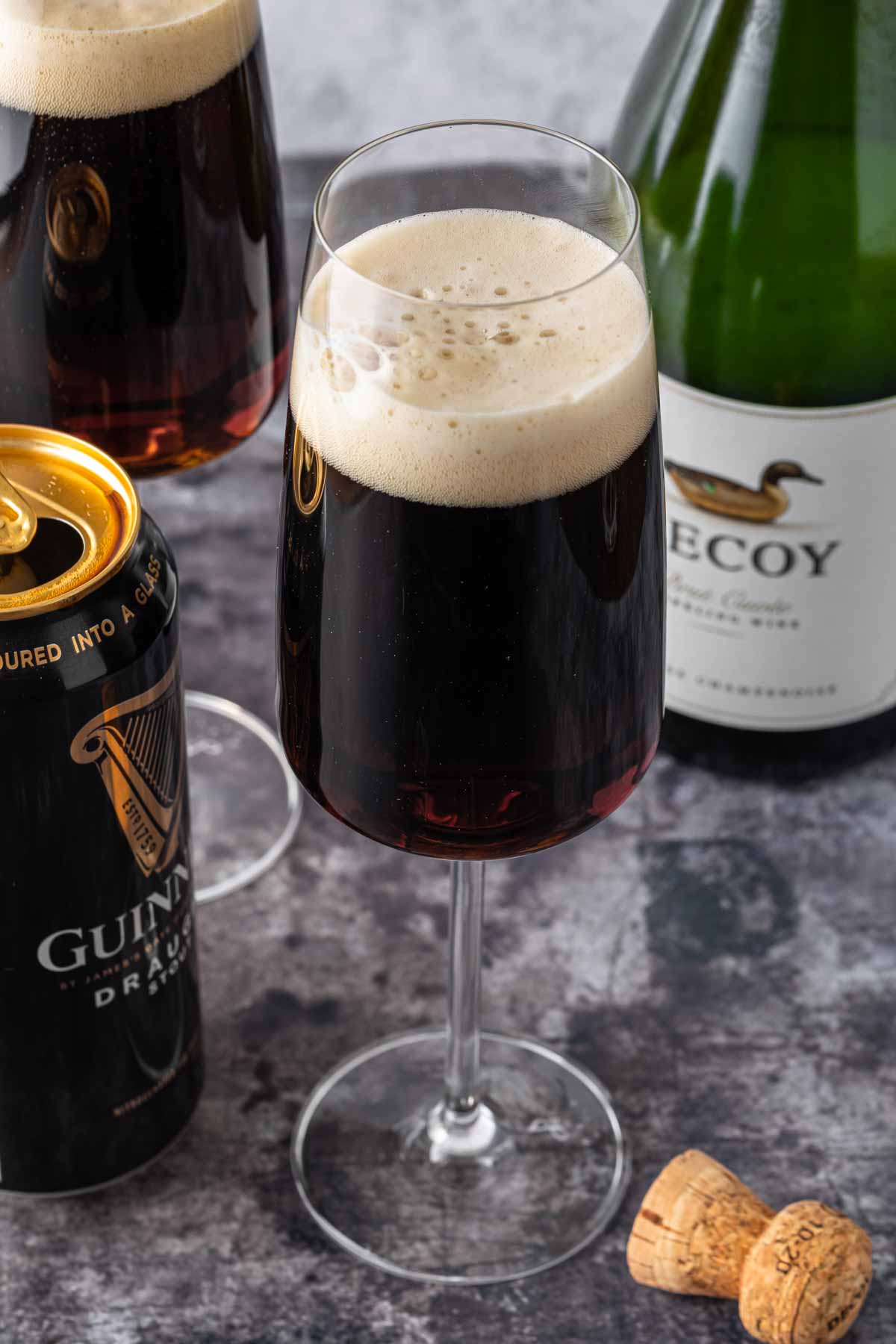 A black velvet cocktail in a champagne glass with a can of Guinness and a bottle of champagne in the background. 
