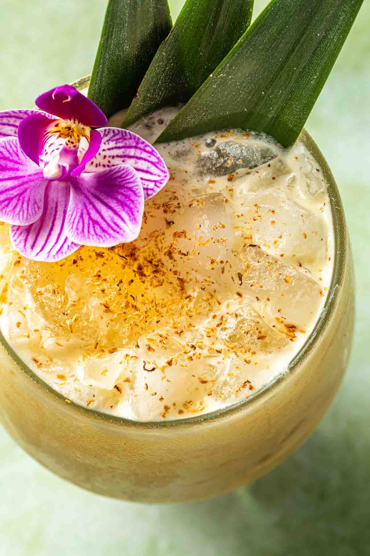Close up of a cocktail showing the grated nutmeg garnish floating on the creamy drink with an orchid flower and pineapple leaves. 