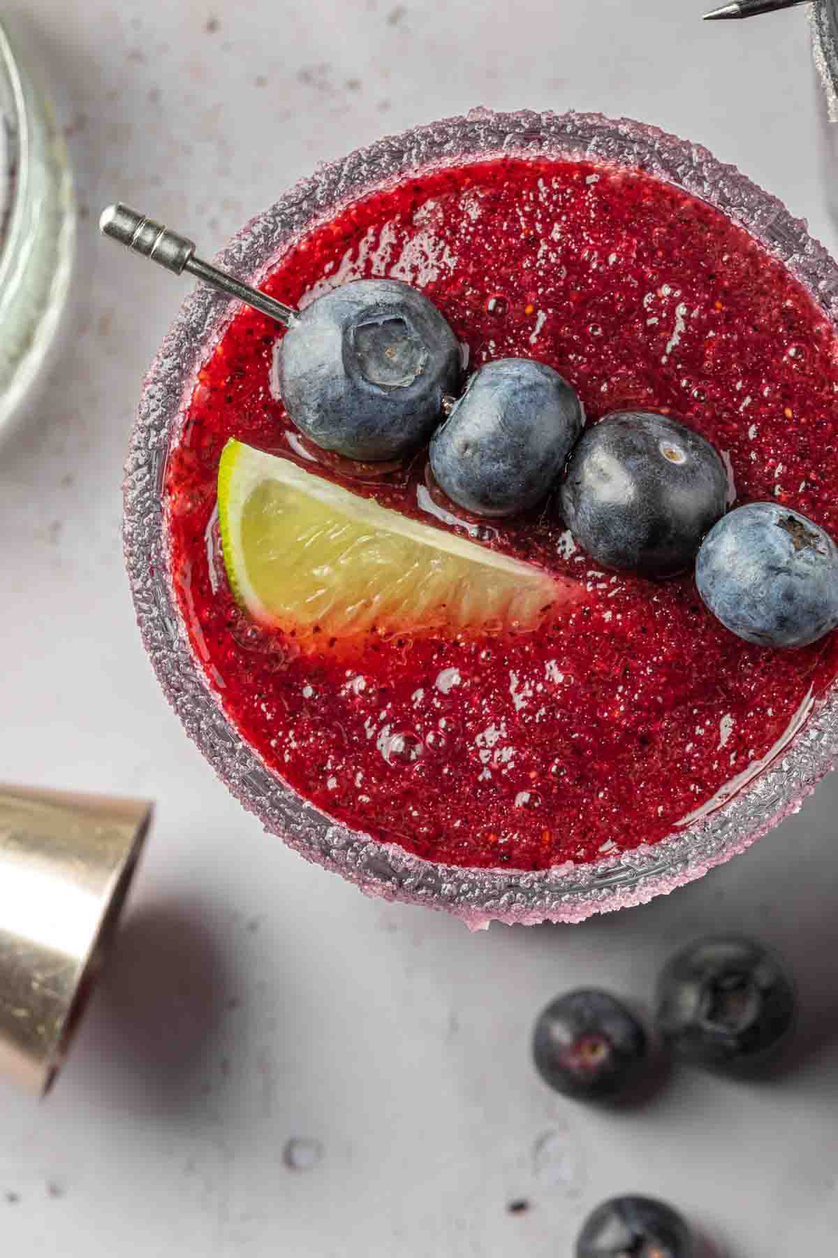 A close up of a blueberry margarita in a sugar rimmed glass, garnished with a cocktail skewer of blueberries and a lime wedge. 