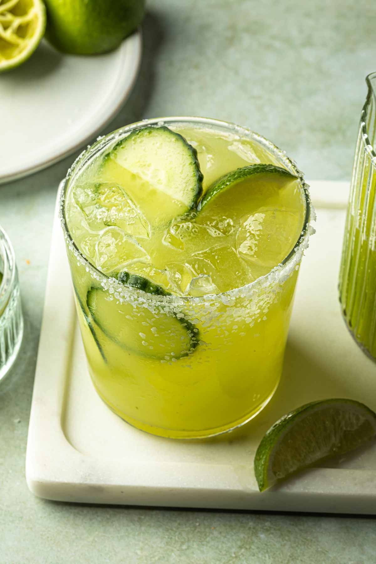 A margarita rimmed with salt on a serving tray next to a small pitcher of cucumber juice. 