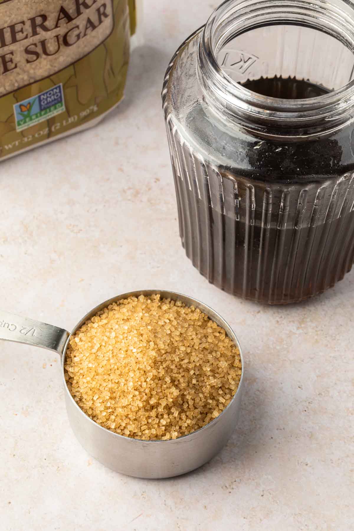 A glass bottle of demerara syrup with a measuring cup of demerara sugar and a bag of the sugar next to it. 