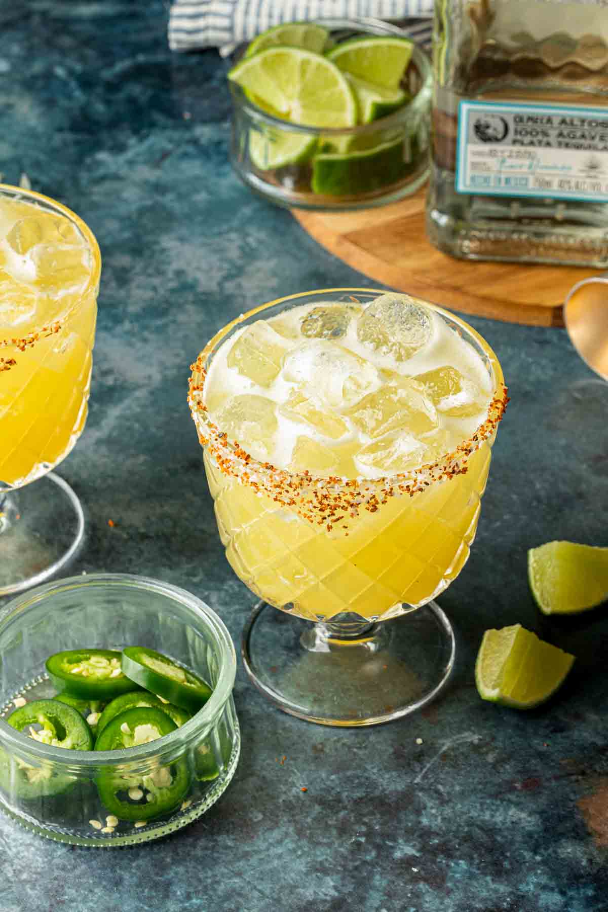 A pineapple margarita in a tajin rimmed glass, surrounded by items used to make the margarita. 