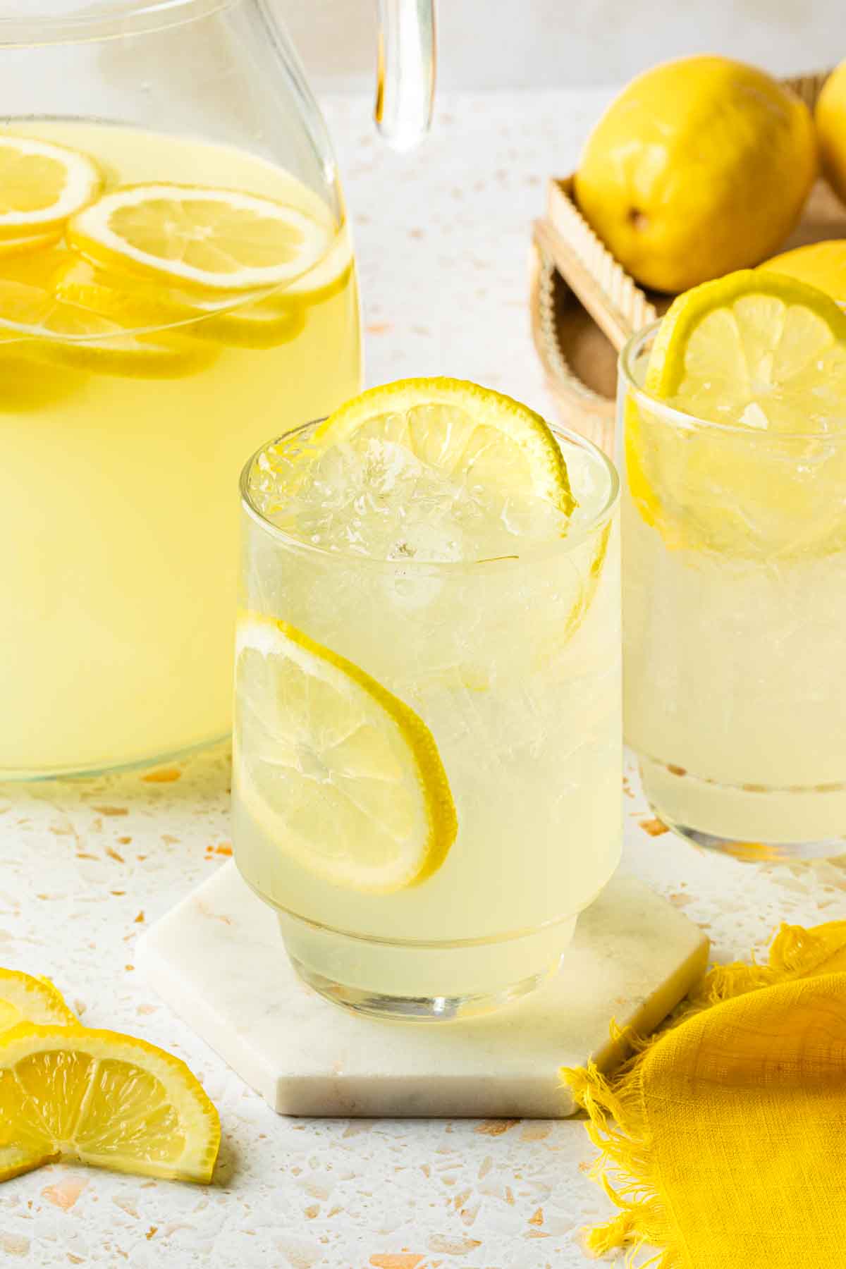 A couple of glasses of lemonade with lemon wheels and a pitcher of tequila lemonade behind them. 
