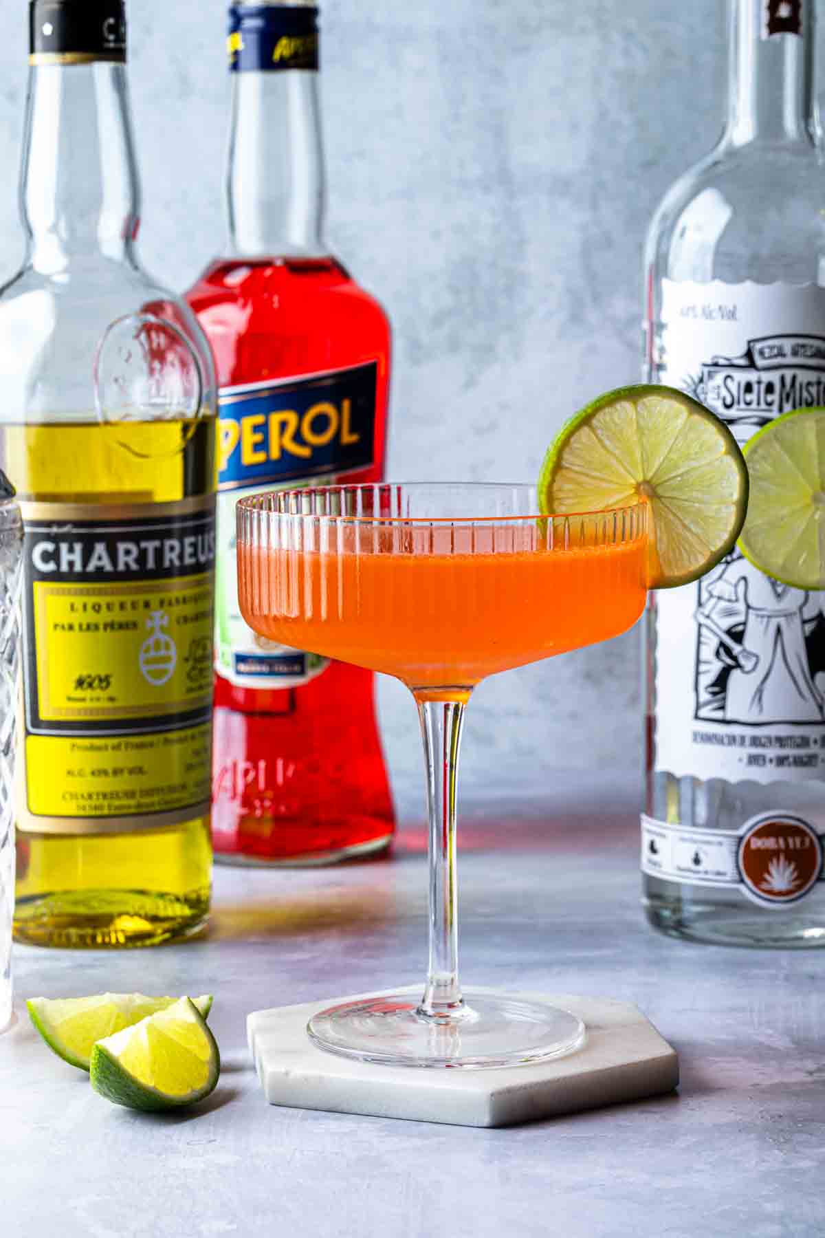 A cocktail in a coupe glass, garnished with a lime wheel. Bottles of yellow chartreuse aperol and mezcal behind the cocktail. 