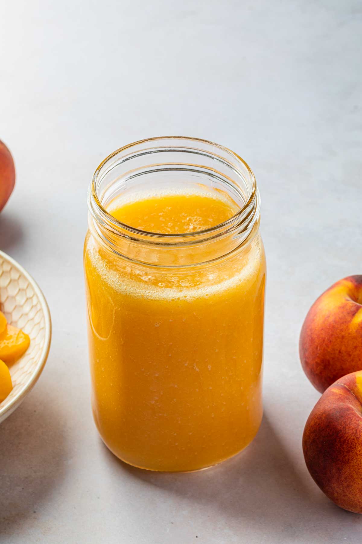A mason jar of peach nectar, next to a  bowl of peaches and a few whole fresh peaches. 