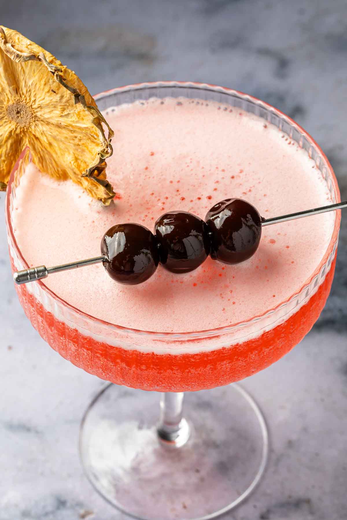 Close up of a cocktail in a coupe glass, garnished with luxardo cherries. 
