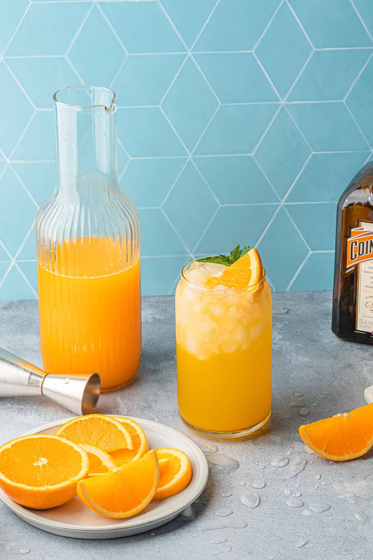 An orange cocktail, a carafe of oj, a bottle of orange liqueur and a plate of oranges with a cocktail jigger. 