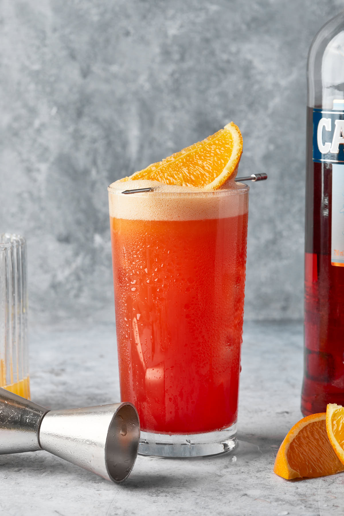 A tall cocktail garnished with an orange wedge, made from orange juice and campari. 