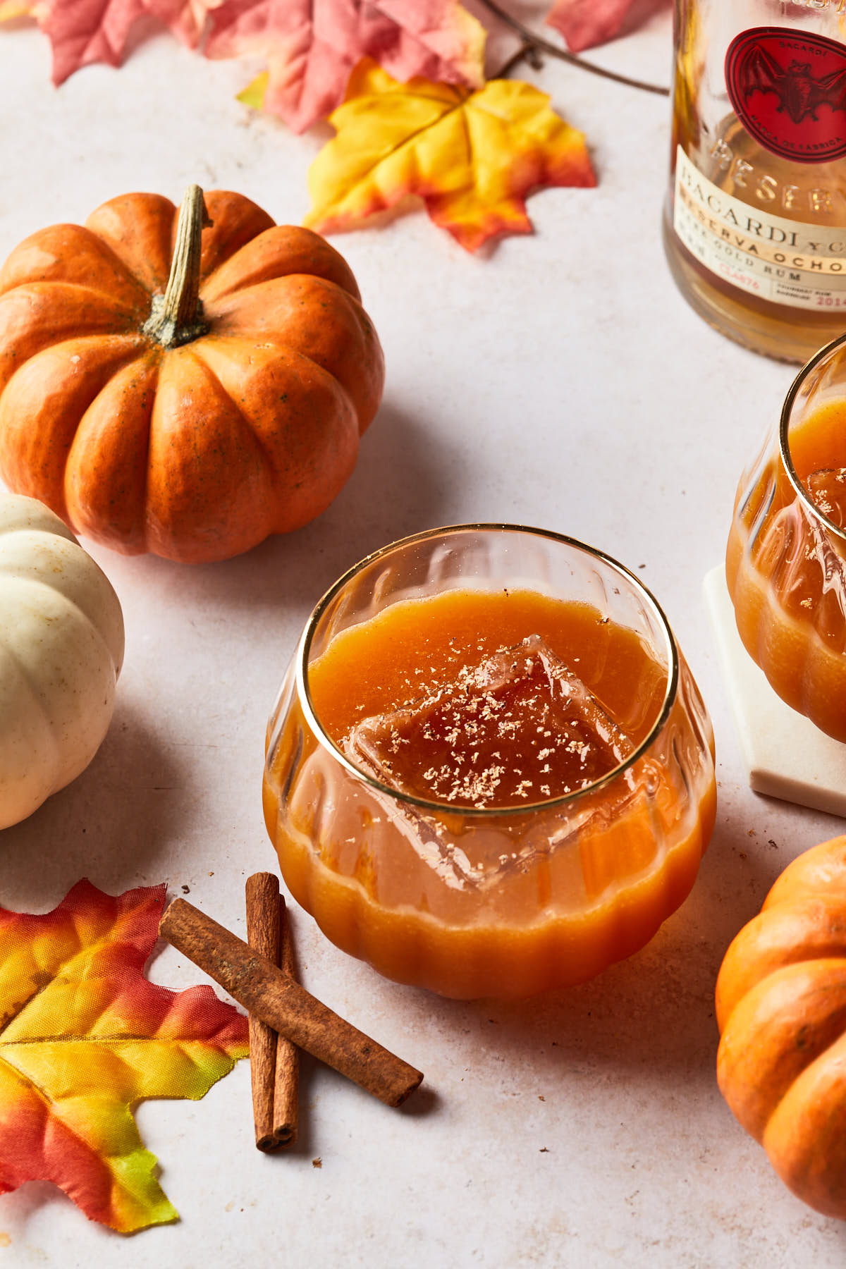 An orange cocktail in a seasonal glass next to cinnamon sticks and fall leaves, and a bottle of dark rum. 
