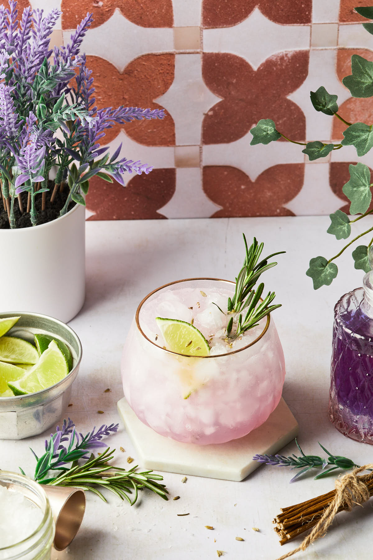 A rosemary lavender margarita with a rosemary sprig garnishing the cocktail surrounded by bunches of herbs, a lavender plant and a bottle of purple simple syrup. 