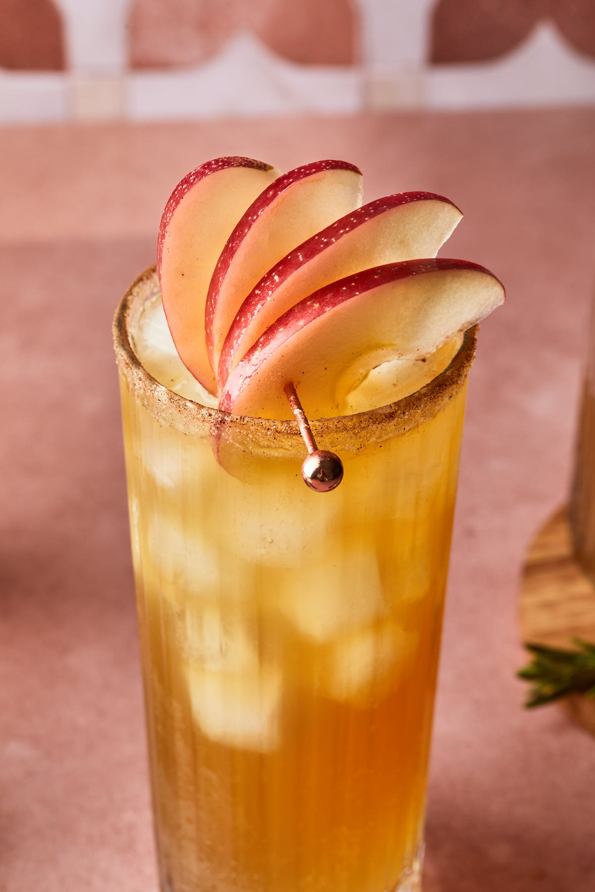 Close up of apple slices on a metal drink skewer garnishing a glass rimmed with cinnamon and sugar. 