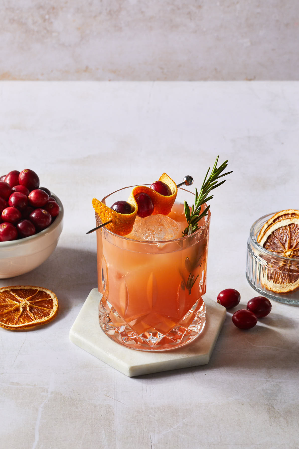 A cranberry orange whiskey sour in a faceted old fashioned glass, garnished with a sprig of rosemary, an orange peel and fresh cranberries. 